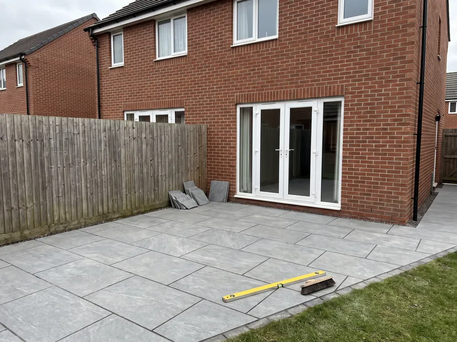 Freshly laid grey porcelain patio behind a red-brick house, with a spirit level and jointing brush resting on the slabs