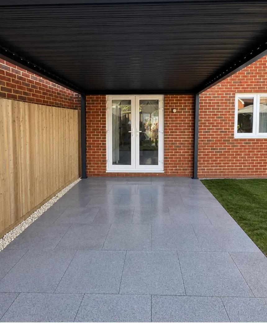 Grey porcelain patio under a black aluminium pergola, edged with white cobbles against a new close-board fence
